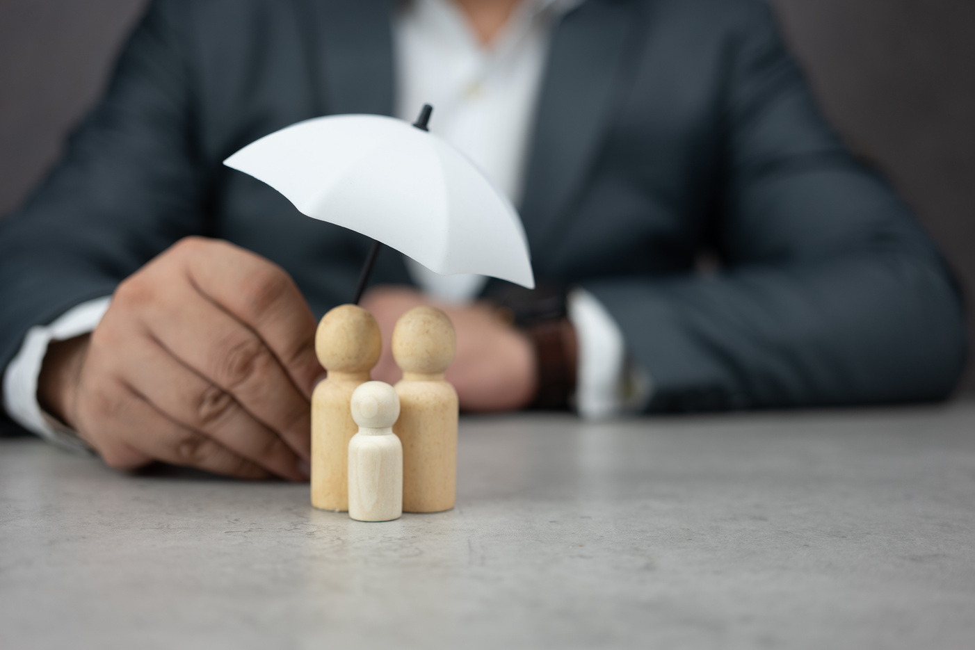 Businessman protecting family of wooden dolls are under a white umbrella, protecting wooden peg dolls.